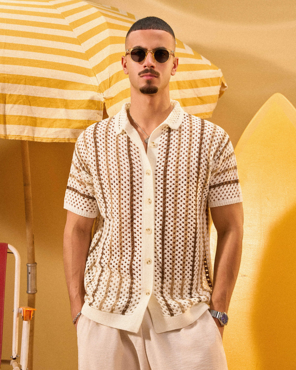 Man wearing a patterned shirt and sunglasses under a yellow and white striped umbrella.