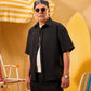 Man wearing sunglasses and a black shirt with a yellow and white striped umbrella and surfboard in the background.