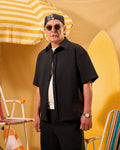 Man wearing sunglasses and a black shirt with a yellow and white striped umbrella and surfboard in the background.