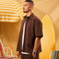 Man in brown outfit standing under a yellow and white striped umbrella with a surfboard in the background.