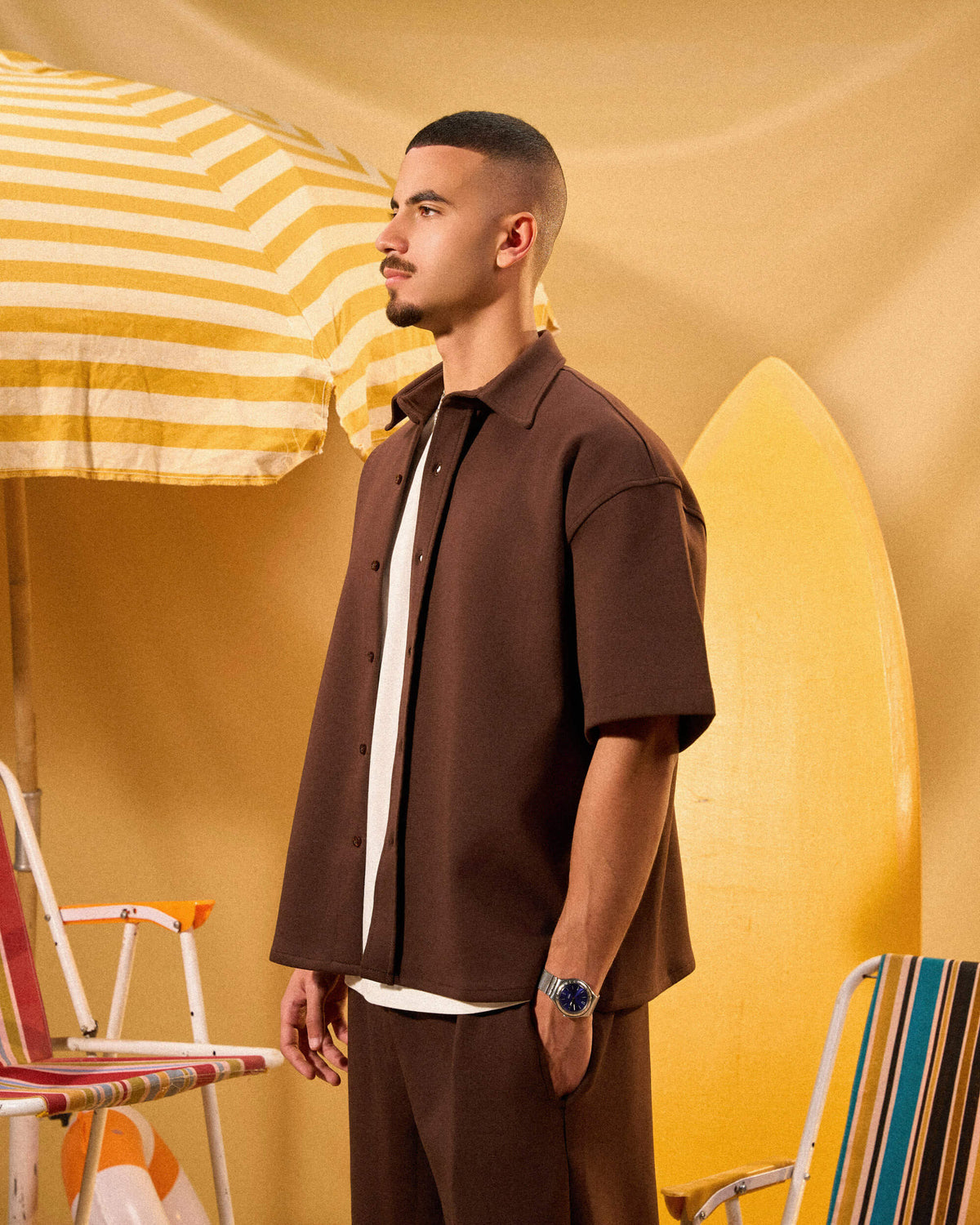 Man in brown outfit standing under a yellow and white striped umbrella with a surfboard in the background.