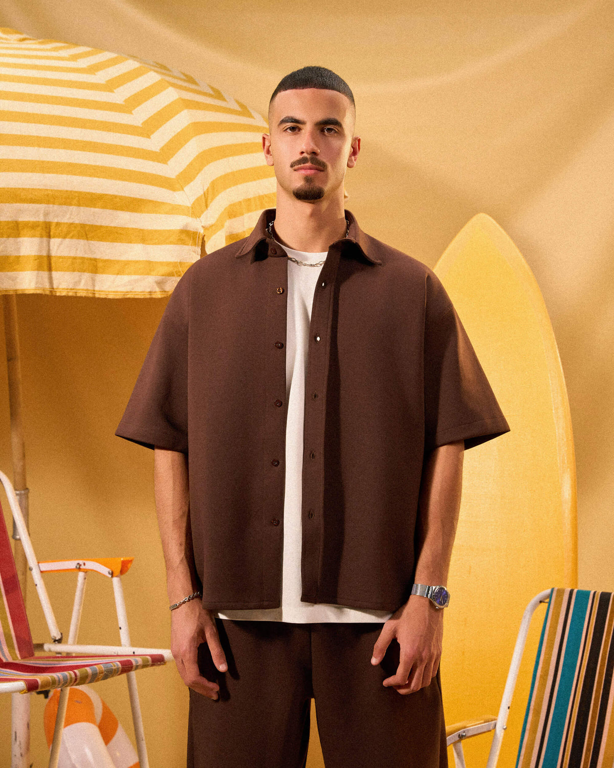 Man wearing a brown shirt and pants standing in front of a yellow and white striped umbrella.