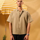 Man wearing a beige shirt and black pants standing next to a yellow and white striped umbrella on a yellow background.