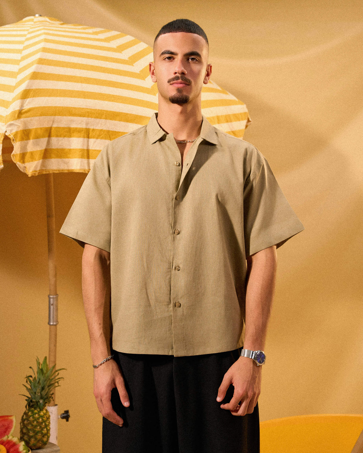 Man wearing a beige shirt and black pants standing next to a yellow and white striped umbrella on a yellow background.