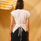 Woman wearing a white top with a tie detail in front of a yellow and white striped umbrella.