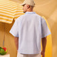 Man wearing a light blue shirt and beige cap standing under a yellow and white striped umbrella with a watermelon on a table.