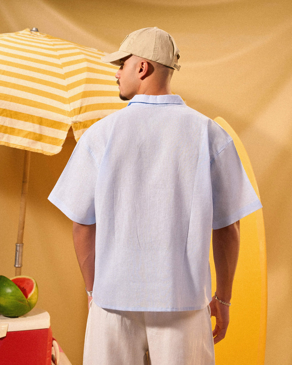 Man wearing a light blue shirt and beige cap standing under a yellow and white striped umbrella with a watermelon on a table.