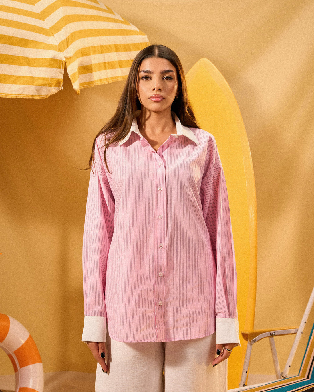 Woman in a pink shirt and white pants standing in a colorful beach-themed setting with chairs and an umbrella.