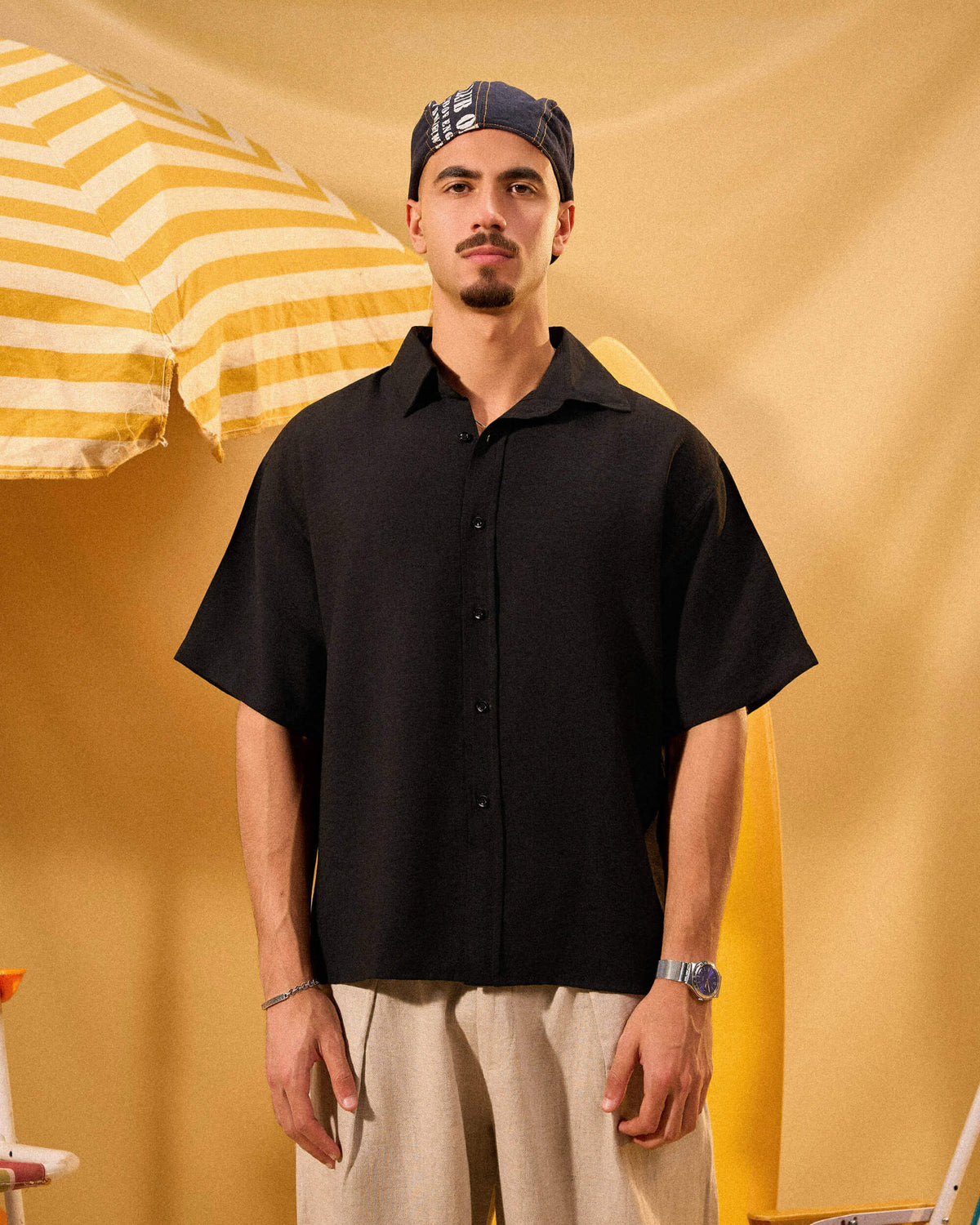 Man wearing a black shirt and beige pants standing against a yellow background with an umbrella.