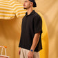 Man wearing a black shirt and cap standing against a yellow background with a striped umbrella.