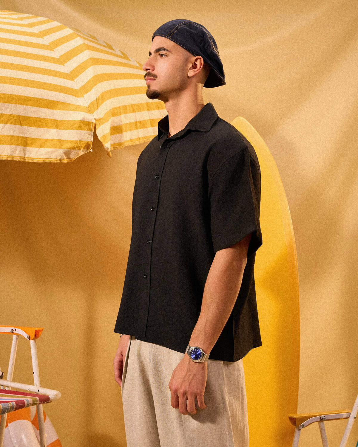 Man wearing a black shirt and cap standing against a yellow background with a striped umbrella.