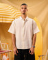 Man wearing a white shirt standing next to a yellow surfboard and striped umbrella on a yellow background