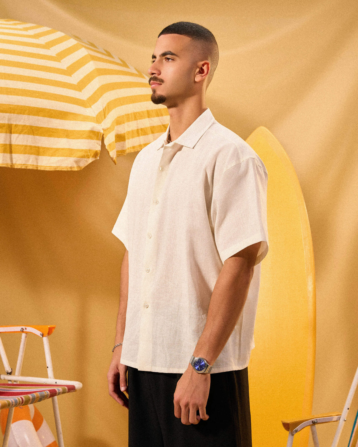 Man in a white shirt standing next to a yellow surfboard and striped umbrella on a yellow background