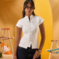 A woman wearing a white short-sleeve mock-neck top with decorative front buttons and dark blue jeans, standing in a stylized yellow beach-themed setting with striped chairs.