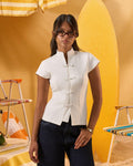 A woman wearing a white short-sleeve mock-neck top with decorative front buttons and dark blue jeans, standing in a stylized yellow beach-themed setting with striped chairs.
