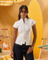 A woman wearing a white short-sleeve mock-neck top with decorative front buttons and dark blue jeans, standing in a stylized yellow beach-themed setting with striped chairs.