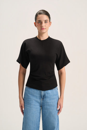 A woman modeling a black short-sleeve crewneck t-shirt paired with light blue jeans, standing against a plain light beige studio background.