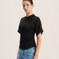 A woman modeling a black short-sleeve mock neck top with gathered side details, paired with light blue jeans, against a plain light gray studio background.