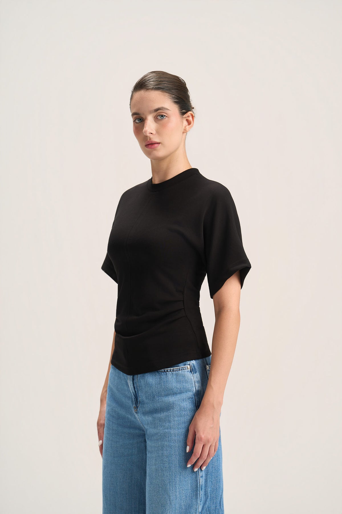 A woman modeling a black short-sleeve mock neck top with gathered side details, paired with light blue jeans, against a plain light gray studio background.