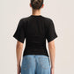 A woman viewed from behind wearing a fitted black short-sleeve t-shirt and light blue jeans against a plain light gray studio background.