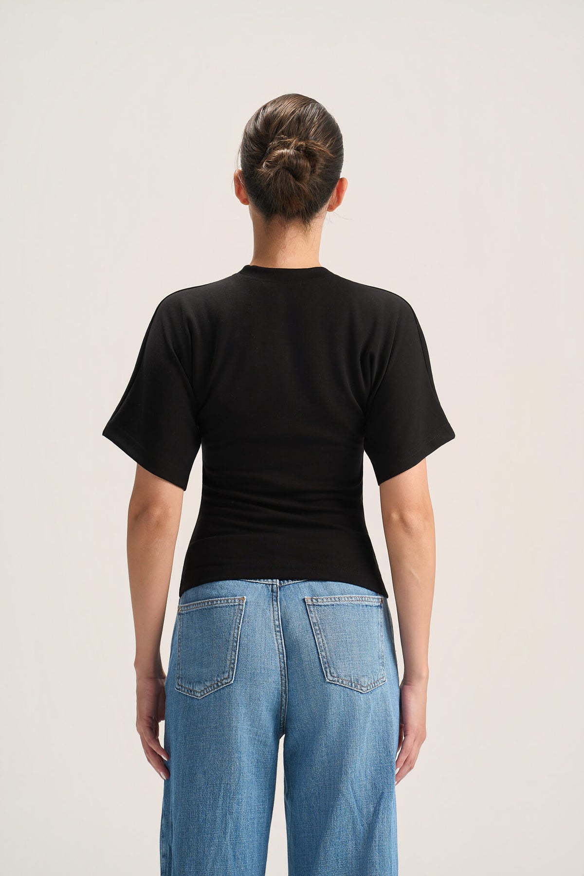 A woman viewed from behind wearing a fitted black short-sleeve t-shirt and light blue jeans against a plain light gray studio background.