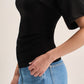 A close-up side view of a person wearing a fitted black short-sleeve top and light blue jeans against a plain light beige background.