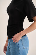 A close-up side view of a person wearing a fitted black short-sleeve top and light blue jeans against a plain light beige background.
