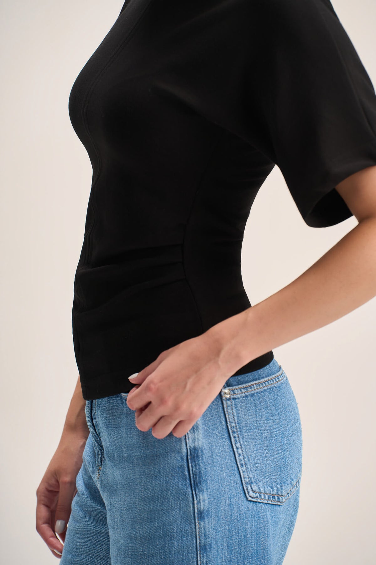A close-up side view of a person wearing a fitted black short-sleeve top and light blue jeans against a plain light beige background.