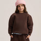 A child model wearing a dark brown crewneck sweatshirt, matching wide-leg sweatpants, a light blue undershirt, a pink knit beanie, and gray slip-on shoes, standing against a neutral light gray studio background.