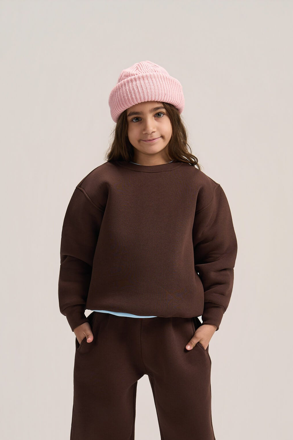 A child model wearing a dark brown crewneck sweatshirt, matching wide-leg sweatpants, a light blue undershirt, a pink knit beanie, and gray slip-on shoes, standing against a neutral light gray studio background.
