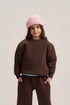 A child model wearing a dark brown crewneck sweatshirt, matching wide-leg sweatpants, a light blue undershirt, a pink knit beanie, and gray slip-on shoes, standing against a neutral light gray studio background.