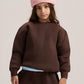 A child model wearing a dark brown crewneck sweatshirt, matching brown sweatpants, a light blue undershirt, and a pink ribbed knit beanie, photographed against a neutral light gray studio background.