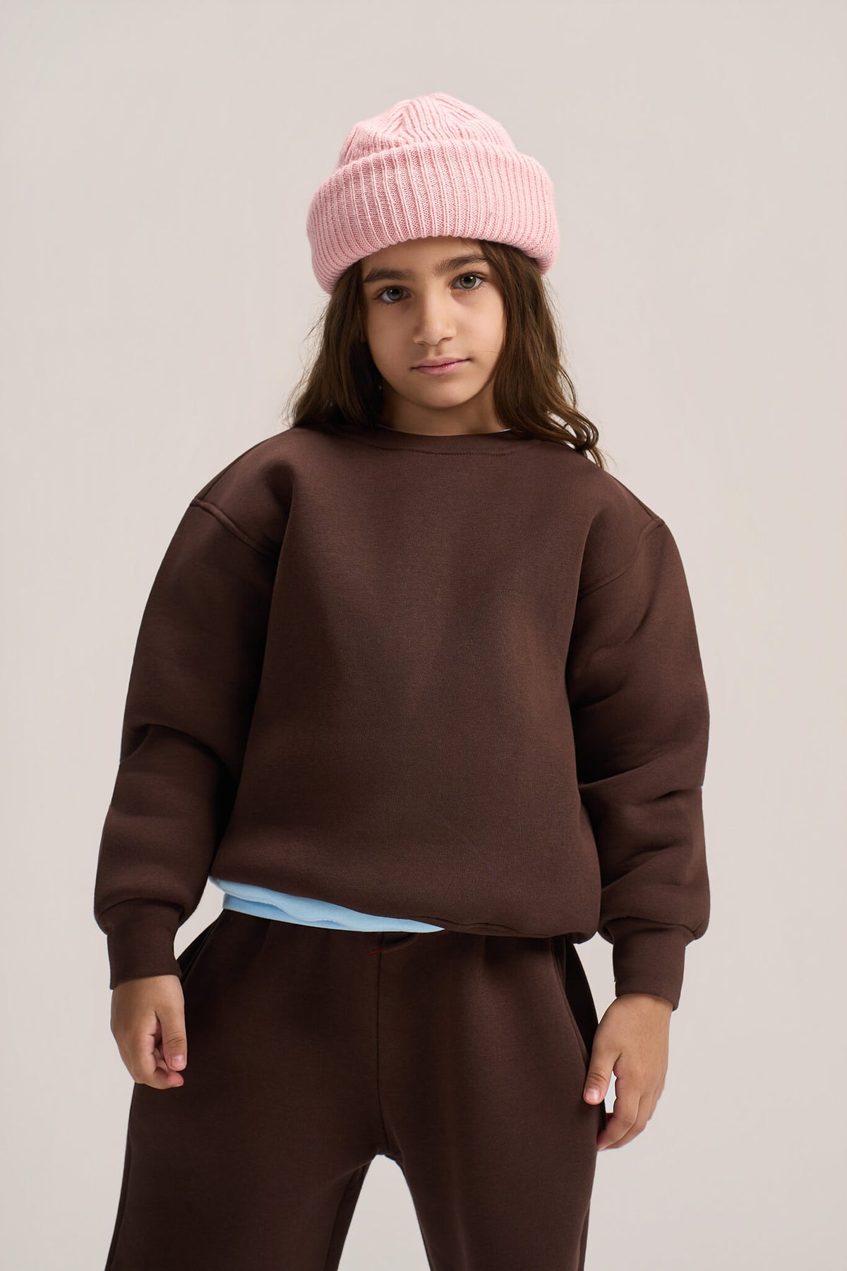 A child model wearing a dark brown crewneck sweatshirt, matching brown sweatpants, a light blue undershirt, and a pink ribbed knit beanie, photographed against a neutral light gray studio background.