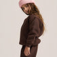 A child model wearing a matching dark brown sweatshirt and wide-leg sweatpants set, styled with a light pink ribbed beanie and brown slip-on shoes, photographed against a solid light gray studio background.