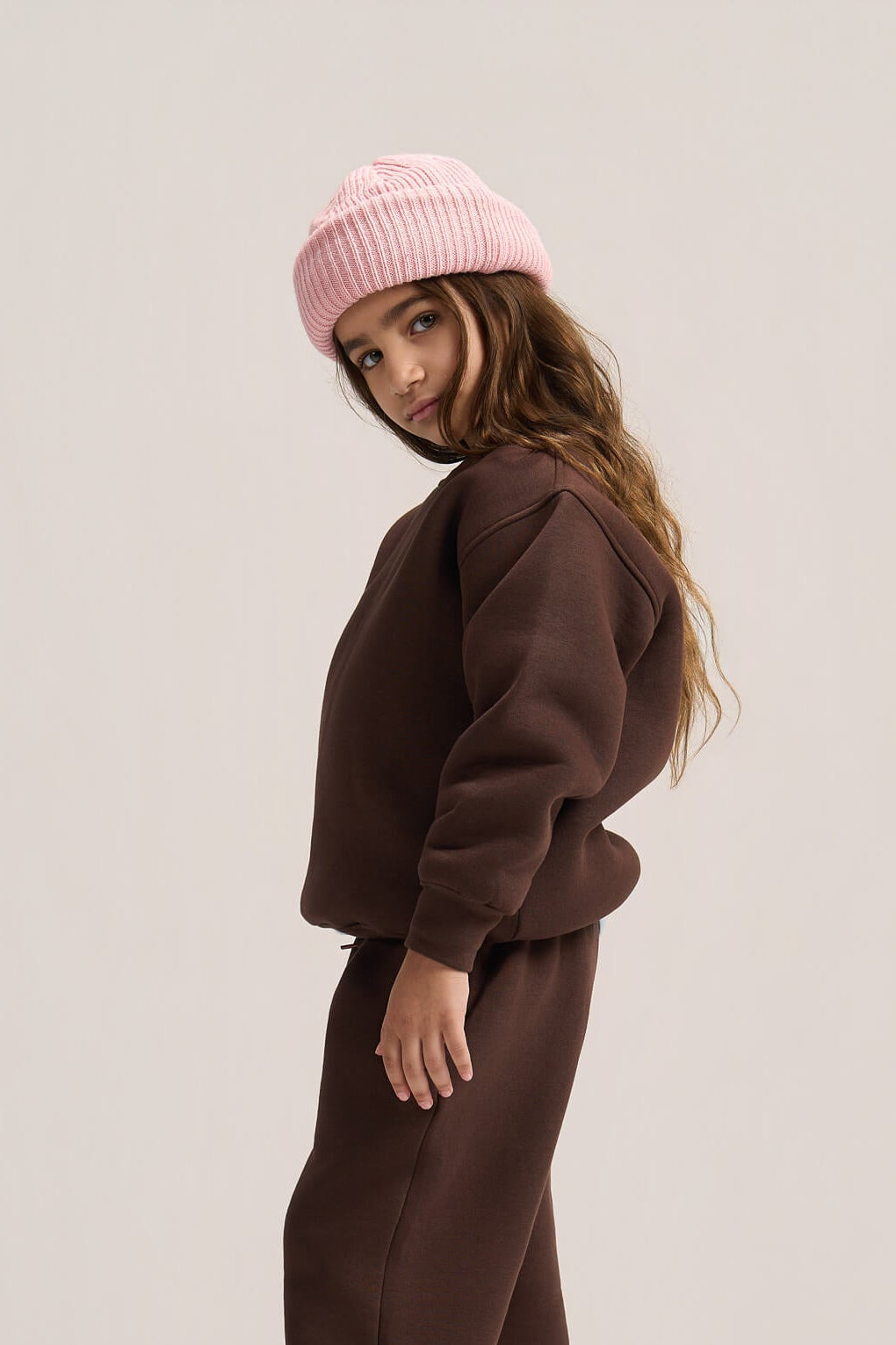 A child model wearing a matching dark brown sweatshirt and wide-leg sweatpants set, styled with a light pink ribbed beanie and brown slip-on shoes, photographed against a solid light gray studio background.