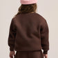 A child shown from the back wearing a dark brown sweatshirt and matching sweatpants, with a light pink ribbed beanie, against a plain light beige studio background.