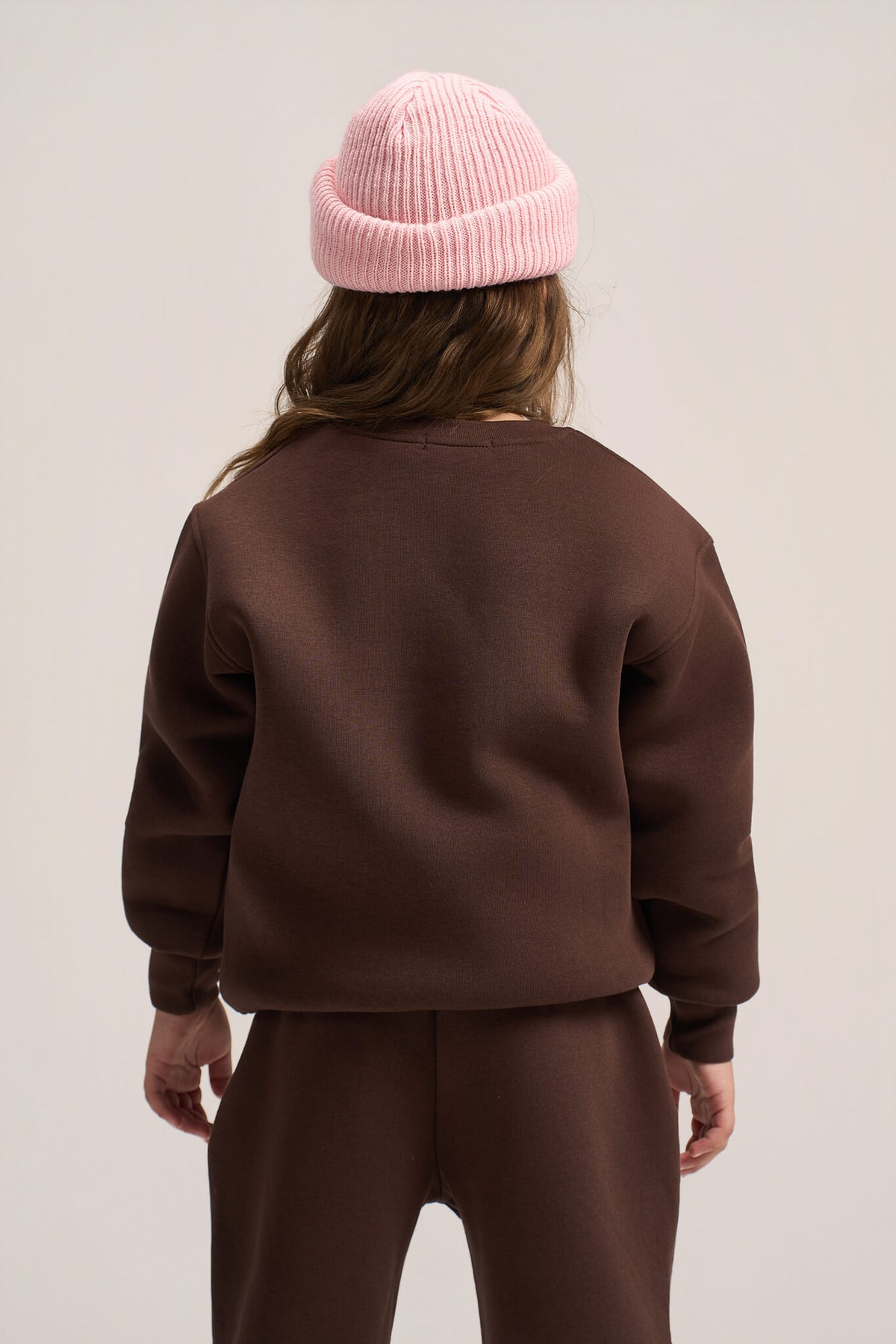 A child shown from the back wearing a dark brown sweatshirt and matching sweatpants, with a light pink ribbed beanie, against a plain light beige studio background.