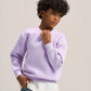 A child model wearing a light purple crewneck sweatshirt over a white t-shirt and blue jeans, standing against a neutral light gray studio background.