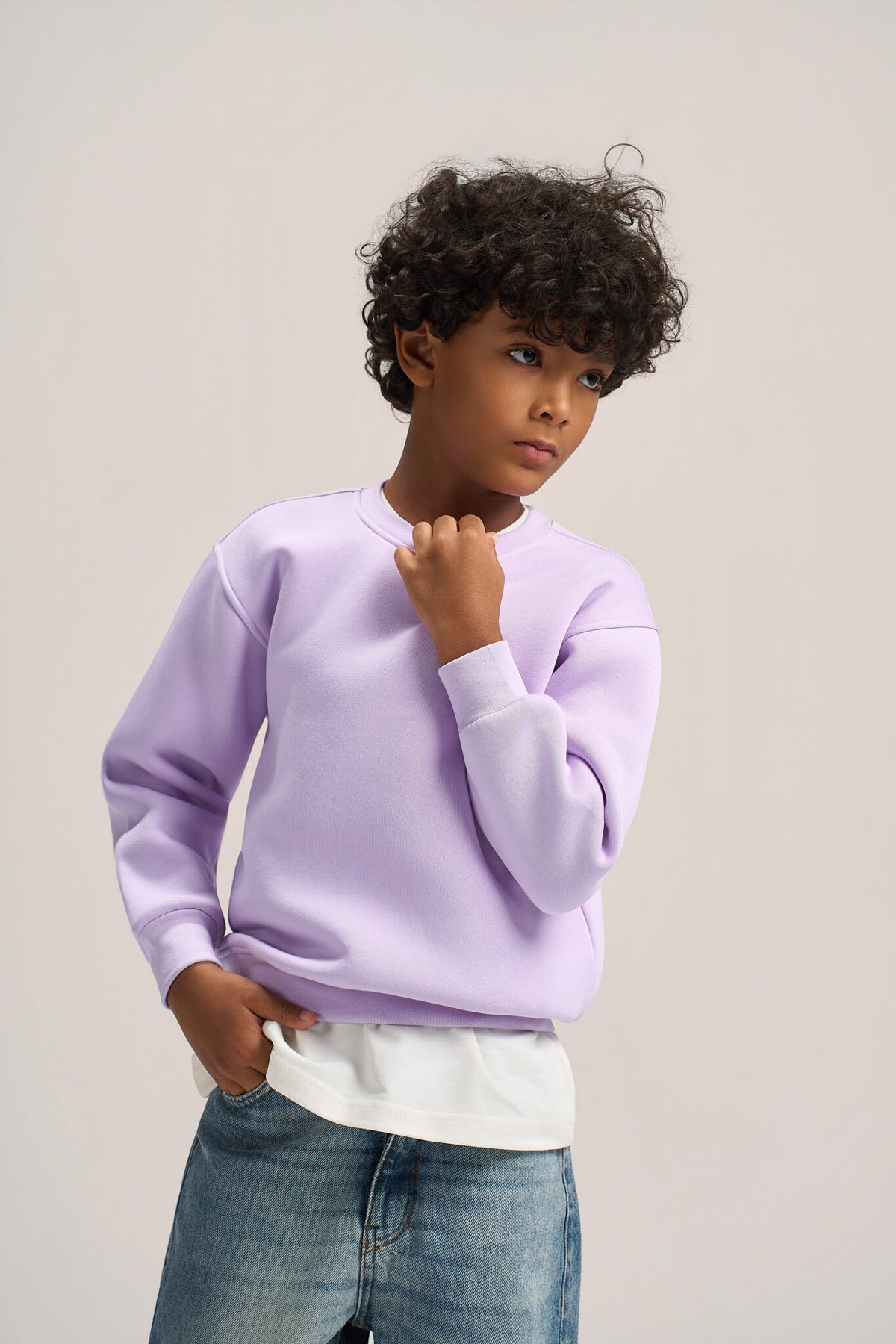 A child model wearing a light purple crewneck sweatshirt over a white t-shirt and blue jeans, standing against a neutral light gray studio background.