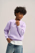 A child model wearing a light purple crewneck sweatshirt over a white t-shirt and blue jeans, standing against a neutral light gray studio background.