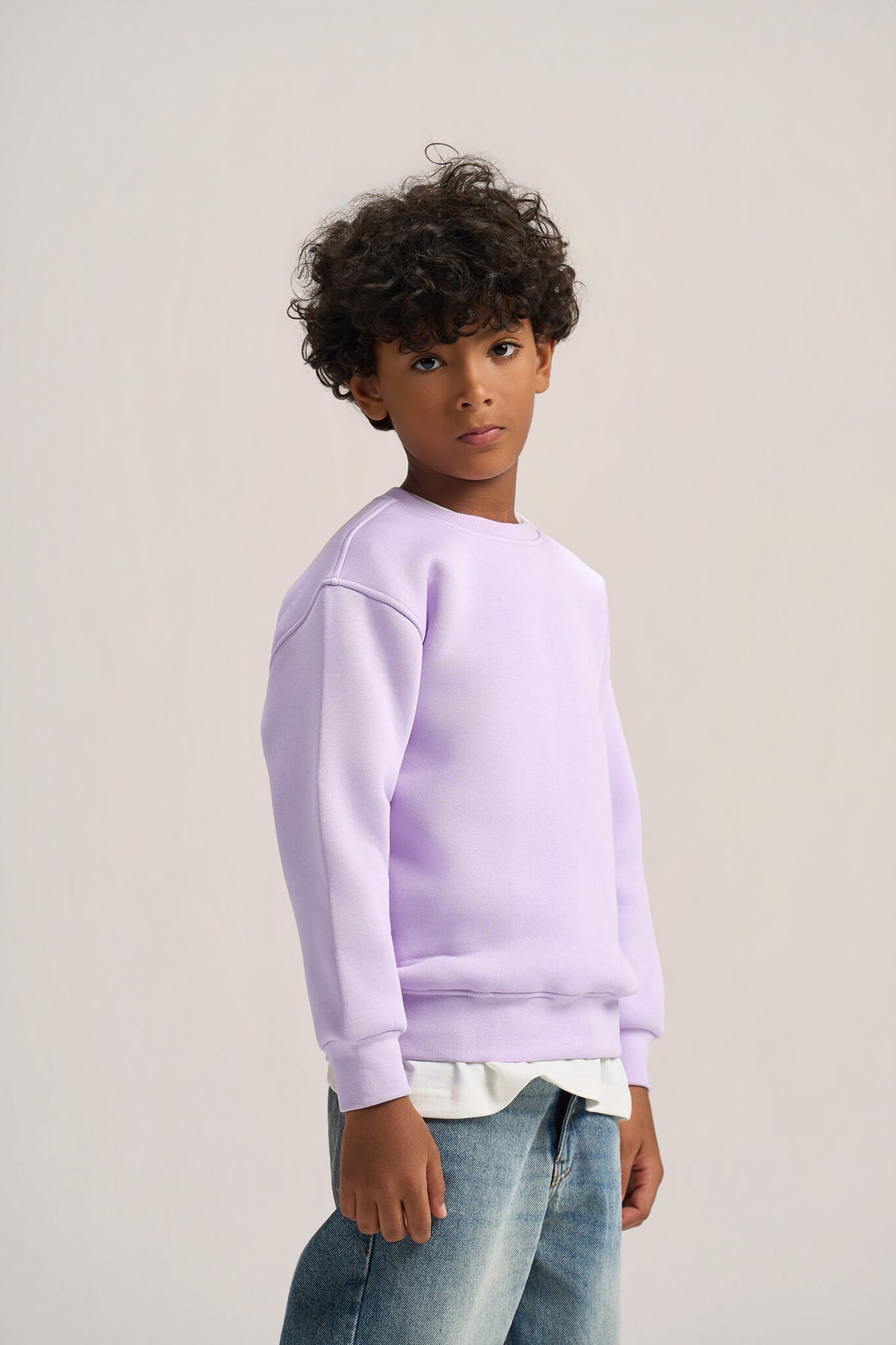 A child model wearing a light purple crewneck sweatshirt, a white undershirt, and blue jeans, photographed against a neutral light gray studio background.