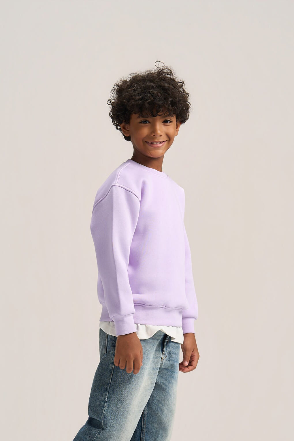 A child model wearing a light purple crewneck sweatshirt, blue jeans, and white sneakers, standing against a solid light gray studio background.