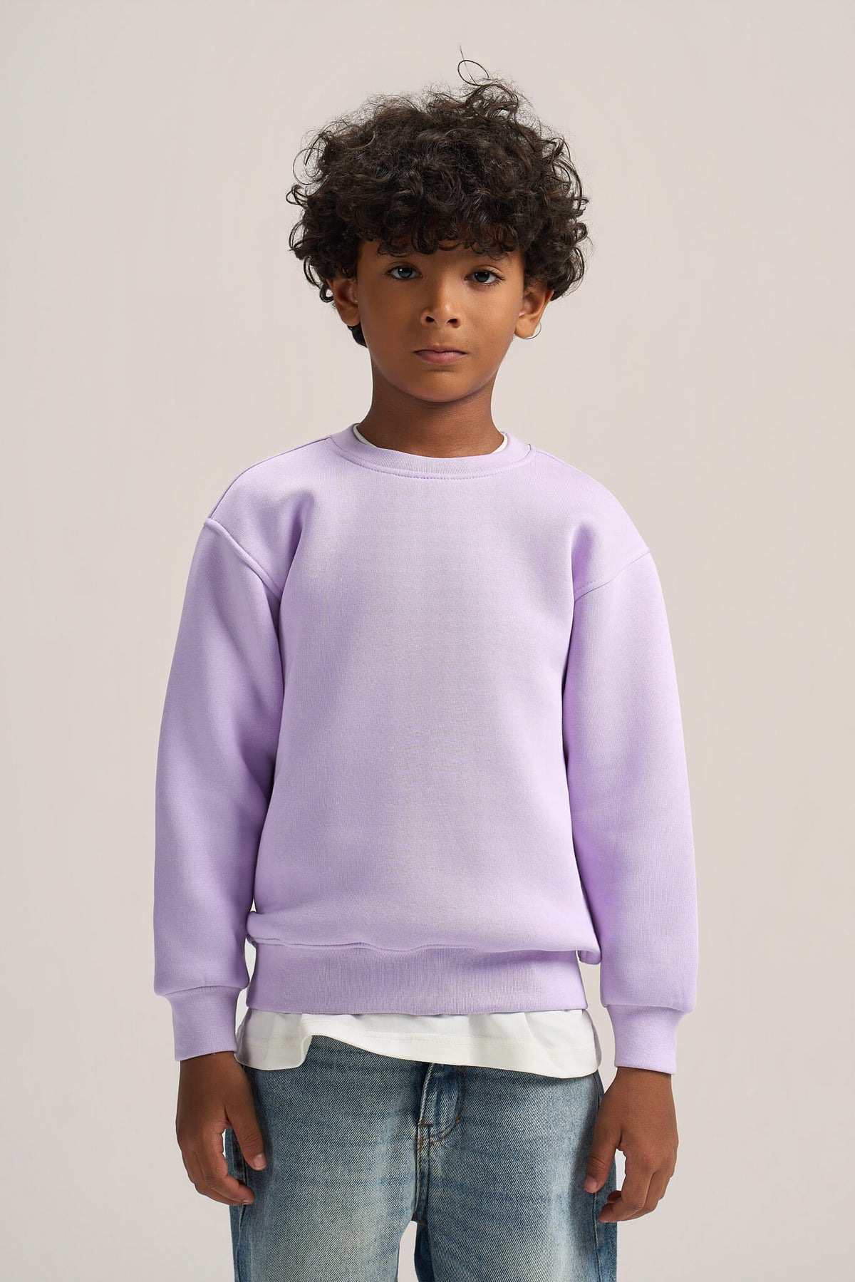 A child model wearing a light purple crewneck sweatshirt, a white t-shirt, and blue jeans, standing against a plain light beige studio background.