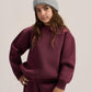 A young girl wearing a matching burgundy sweatshirt and sweatpants set with a light gray knit beanie, posing against a neutral light gray background.