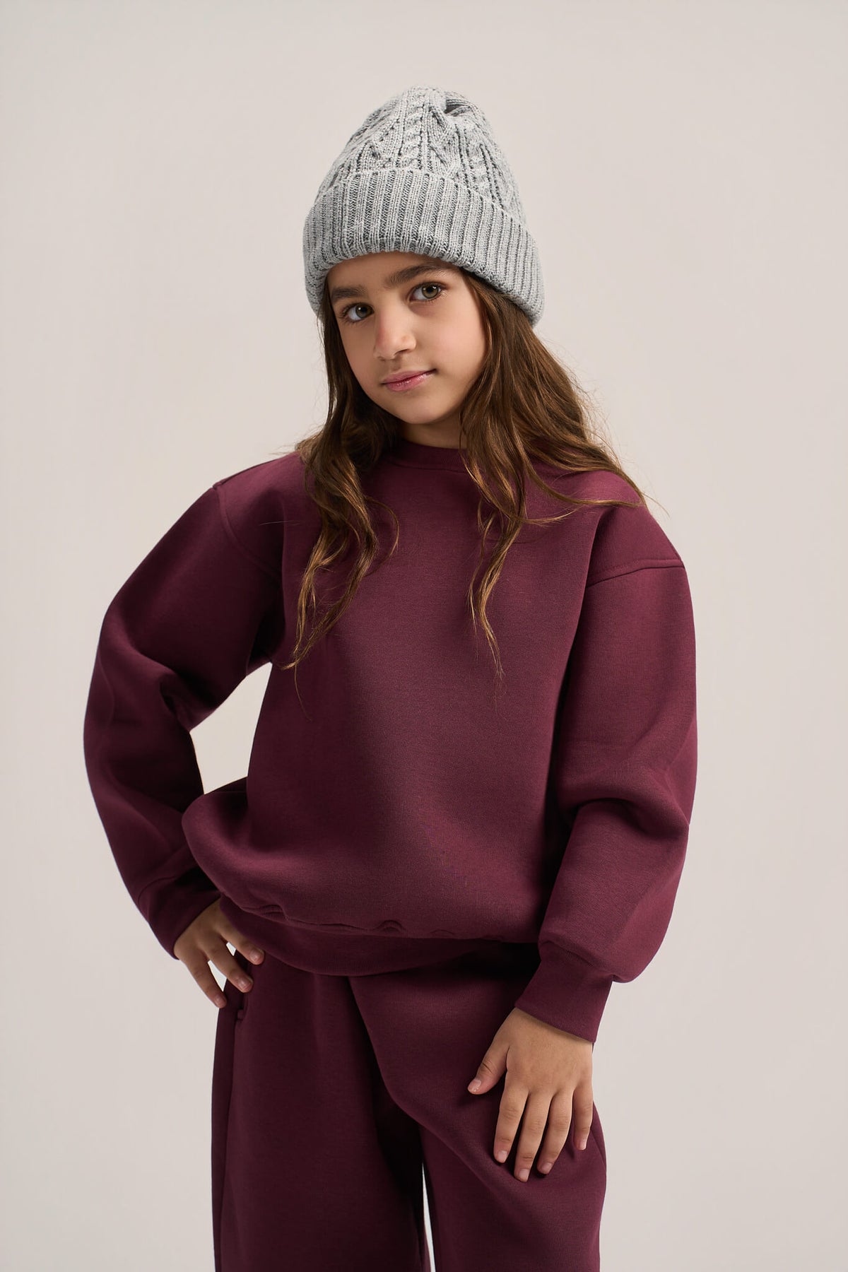 A young girl wearing a matching burgundy sweatshirt and sweatpants set with a light gray knit beanie, posing against a neutral light gray background.