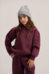 A young girl wearing a matching burgundy sweatshirt and sweatpants set with a light gray knit beanie, posing against a neutral light gray background.