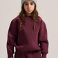 A young girl wearing a matching burgundy sweatshirt and sweatpants set with a light gray knit beanie, posing against a solid light beige background.