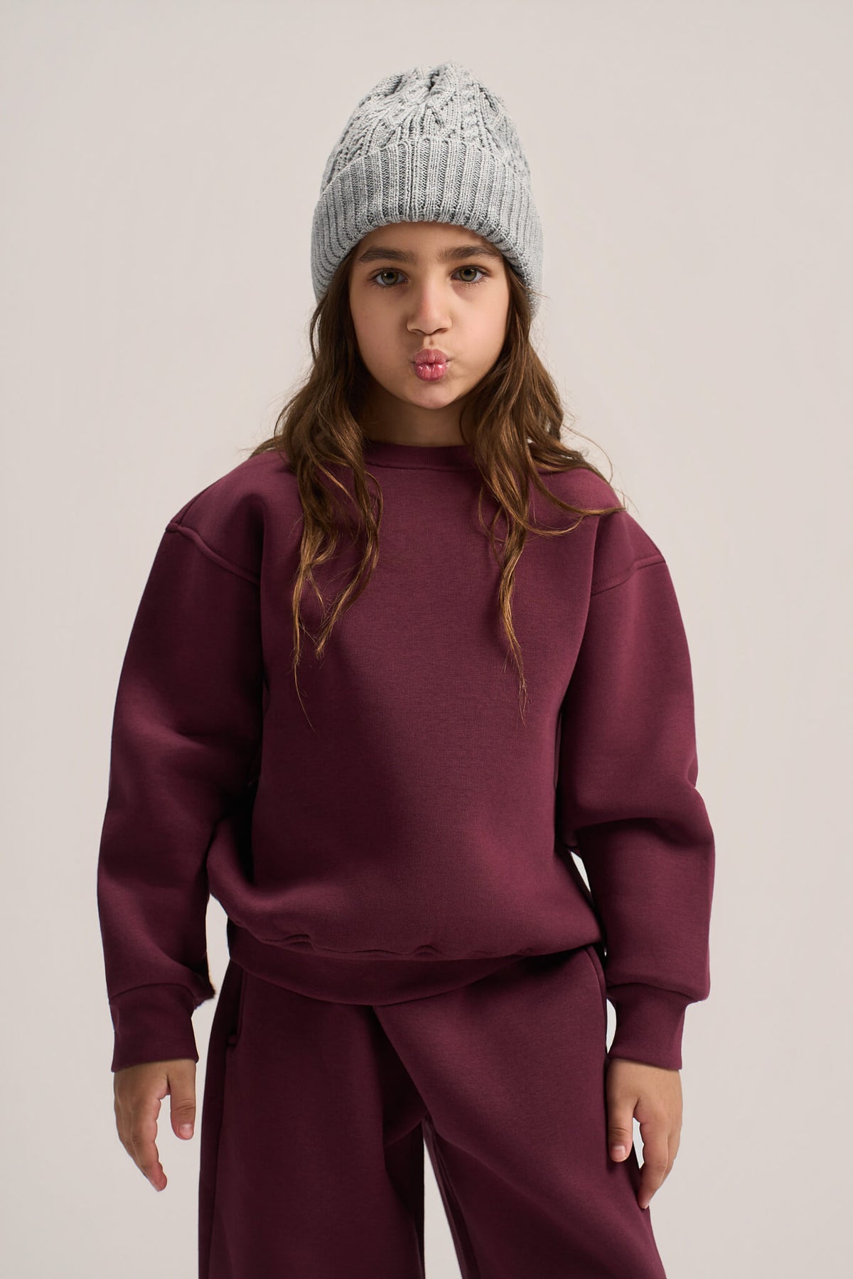A young girl wearing a matching burgundy sweatshirt and sweatpants set with a light gray knit beanie, posing against a solid light beige background.