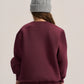 A person shown from the back wearing a dusty burgundy crewneck sweatshirt and matching sweatpants, paired with a light gray cable-knit beanie, against a plain light beige studio background.