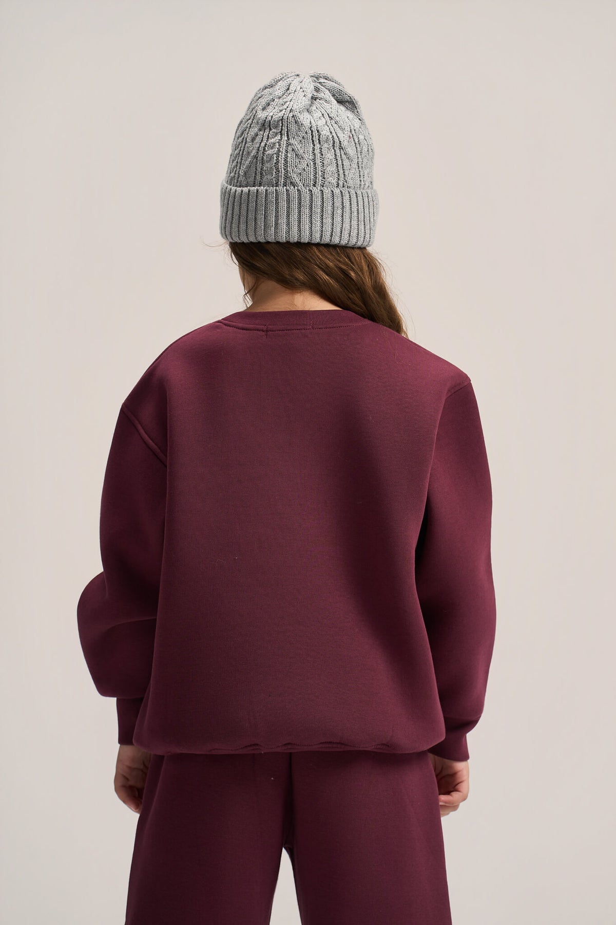 A person shown from the back wearing a dusty burgundy crewneck sweatshirt and matching sweatpants, paired with a light gray cable-knit beanie, against a plain light beige studio background.